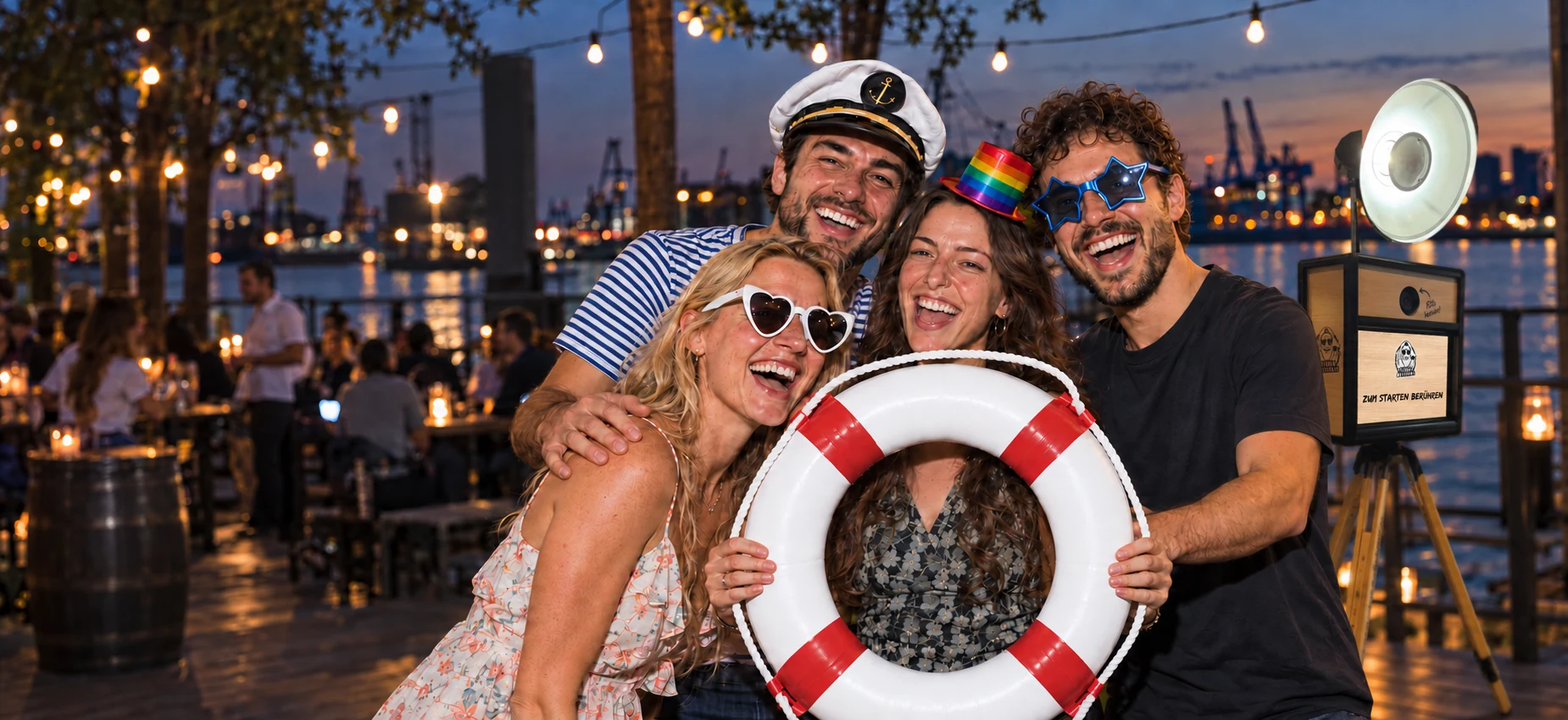 Vier fröhliche Gäste mit Party-Accessoires und Rettungsring, dahinter die Fotobox Classic am Hamburger Hafen